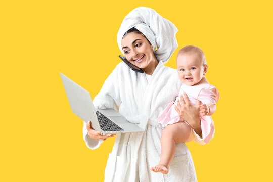 Young business mother in bathrobe with cute little baby and laptop talking by phone on yellow background - Powered by Adobe