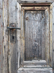 Close-up of an old wooden door with weathered texture, visible grain, peeling paint, and a rusty metal handle with a keyhole, showing signs of age and decay.