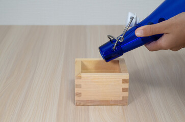 Hand pouring liquid from blue glass bottle into wooden masu cup
