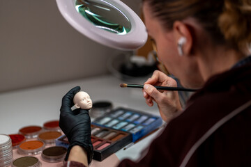 Artist in black gloves painting ball jointed doll face with brush and pigments under magnifying lamp creating custom details using pastel palette in workspace