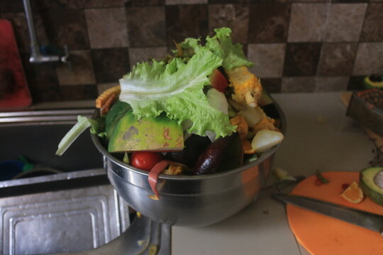 A close-up shot of a stainless steel bowl filled with various organic food scraps and kitchen waste, including lettuce leaves, corn cobs, tomato ends, watermelon rinds, and other vegetable and fruit p