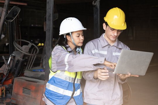Factory engineers review safety plan on laptop inside industrial workshop, focused teamwork under hard hat and vest near forklift with calm confidence and dust haze for maintenance check