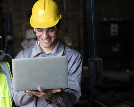 Smiling engineers in hard hat review laptop at factory floor with industrial machinery in background, confident teamwork and focus on safety and quality control drive productive workflow