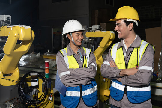 Smiling engineers safety helmet and reflective vest talk beside yellow robotic arm inside factory, teamwork energy industrial setting with modern automation and focused collaboration workshop light