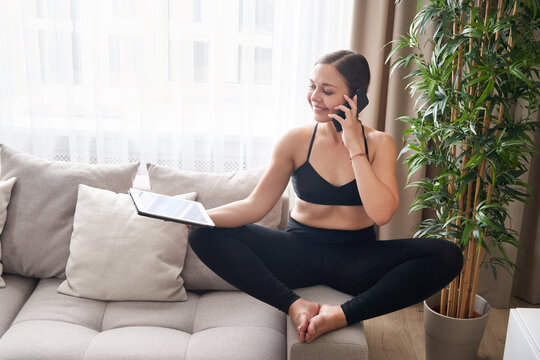 Young woman sitting cross-legged on couch in lotus pose using tablet and phone at home in living room