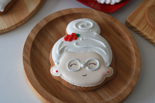 A festive Christmas display with various decorated cookies (Santa, Mrs. Claus, gingerbread man) on wooden boards and a heart-shaped plate, surrounded by holiday decor. - Powered by Adobe