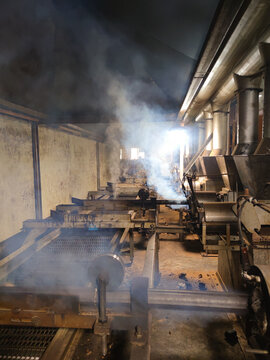 Smoky factory interior with interconnected metal pipes, conveyor systems, and heavy machinery working in a warm dim light, showing raw production atmosphere
