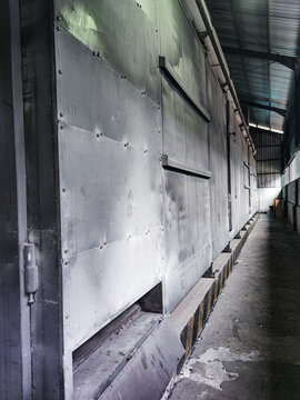 Long factory corridor with a massive metal chamber wall covered in dust and gritty textures. Dim industrial light reveals bolts, panels, and rugged surfaces typical of heavy manufacturing environments
