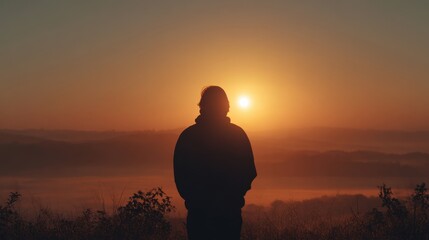 Fototapeta premium Silhouette of a person standing on a hill watching the sunrise over a misty valley