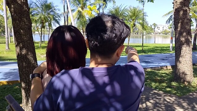 Back view of a couple sitting on a bench in a sunny park. The man points toward a lake while embracing the woman under tree shadows.