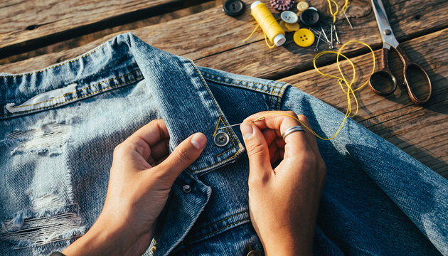 Hands sewing a yellow button onto a blue denim jacket on a rustic wooden surface.