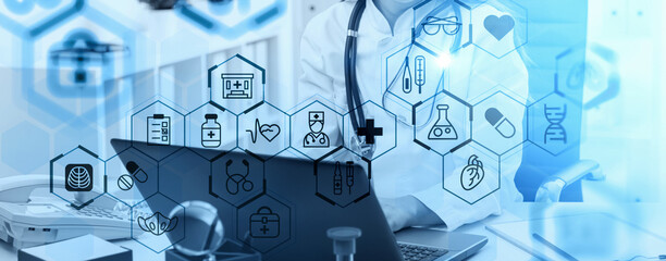 Doctor working on laptop with overlay of medical icons in blue transparent graphic style on hospital office background, concept of digital healthcare innovation