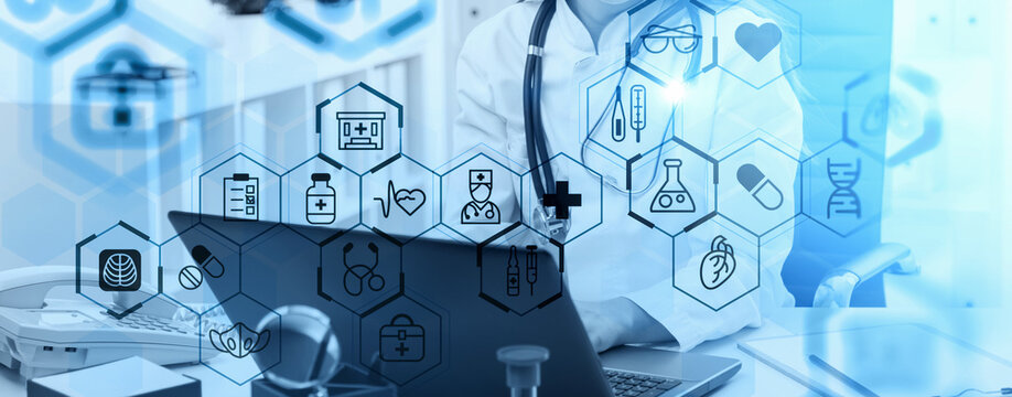 Doctor working on laptop with overlay of medical icons in blue transparent graphic style on hospital office background, concept of digital healthcare innovation