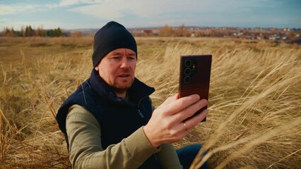 A Man Sitting in Golden Grassland Holds a Smartphone While Gazing Thoughtfully into the Distance Beneath a Bright Blue Sky with Wispy Clouds and Rolling Hills in the Background