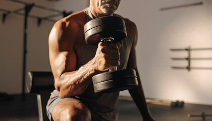 Close up of a senior man with a muscular physique doing a bicep curl with a heavy dumbbell during a strength training session in the gym