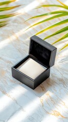 An open black jewelry box on a marble surface with palm leaves, lit by sunlight. The box is empty and ready for a precious item.