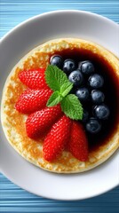 A close-up, top-down view of a golden-brown pancake served on a white plate. The pancake is topped with fresh strawberries, blueberries, and a sprig of mint, wi