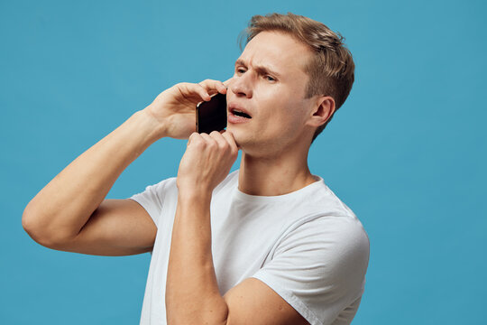Modern young man with short hair making a concerned face while talking on a smartphone isolated on blue background. Studio portrait of adult male with expressive emotions, casual white t-shirt, and