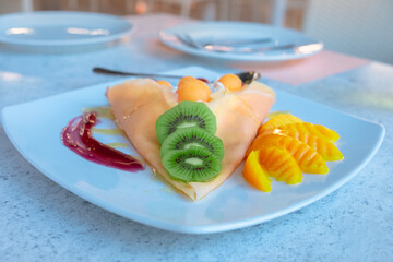 Cold crepe topped with kiwi, mango and cantaloupe sit on a white plate with morning sunlight streaming through.
