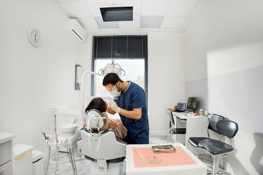 Middle Eastern male dentist treating middle-aged female patient in dental office, dentist wearing protective mask and gloves, patient reclining in dental chair during examination