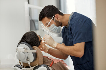 Middle aged Caucasian woman sitting in dental chair, receiving oral examination from young adult male dentist wearing protective mask and gloves in modern dental office