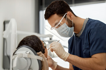 Young adult male dentist wearing protective mask and face shield treating patient in dental office, using dental instruments while focused on dental procedure, patient reclining in dental chair