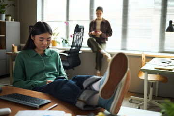 Young adult Asian woman sitting at desk with legs resting on tabletop using smartphone while another young adult Asian woman standing in background near window, holding mobile device in modern office