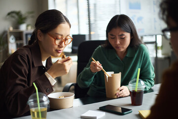 Young Asian businesswomen sitting at office table eating lunch from takeaway containers, drinking beverages with straws, working together during break, smartphone on desk