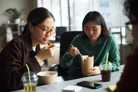 Young Asian businesswomen sitting at office table eating lunch from takeaway containers, drinking beverages with straws, working together during break, smartphone on desk