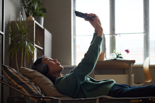 Young adult Asian woman lying on couch wearing headphones holding smartphone above face, relaxing in modern office setting with sunlight streaming through window in background