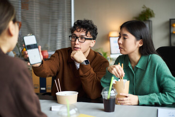 Young Asian man showing smartphone screen to young Asian woman and another young Asian woman, while sitting at office table eating takeout food and drinking beverages during work meeting