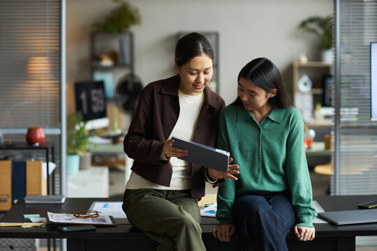 Two young adult Asian women collaborating in modern office, standing and sitting on desk while discussing information displayed on digital tablet, engaging in teamwork and communication