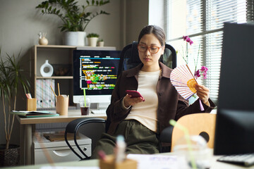 Young adult Asian woman sitting at office desk using smartphone and holding paper fan, computer monitor with code on screen in background, working in modern workspace