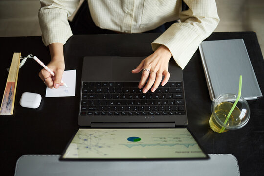 Young Asian woman working at desk using laptop, writing notes on paper with pen, hand visible next to digital tablet and drink, demonstrating multitasking in modern office environment