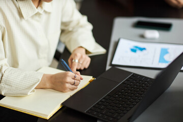 Young Asian woman writing in notebook while working at desk with laptop and digital tablet, focusing on business tasks in modern office environment, hands visible