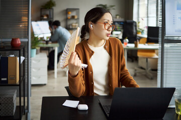 Young adult Asian woman sitting at desk working on laptop, holding handheld fan and talking,...