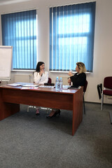 Women discuss charts and documents while sitting at a desk in a bright office with water and notebooks.