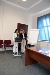 Two women in an office stand by a flipchart, reviewing documents and discussing data during a work presentation.