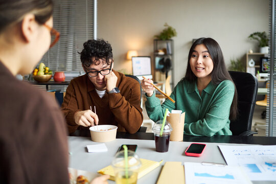 Young Asian woman gesturing with chopsticks while talking to young Asian man eating lunch with chopsticks, both sitting at office desk with documents and drinks, coworker standing nearby