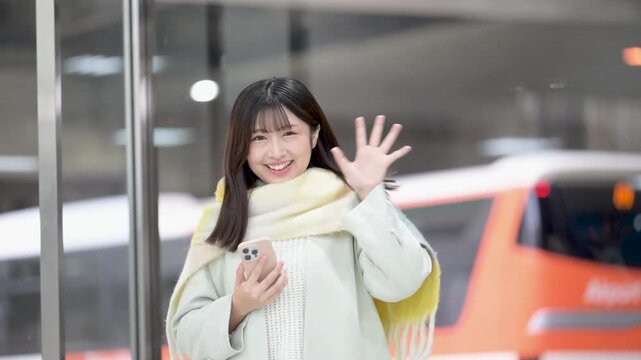 冬の年末年始の帰省、駅のホームやバスターミナルなどでスマホを持つかわいいストールを巻いた女性、手を振るバイバイのスロー動画