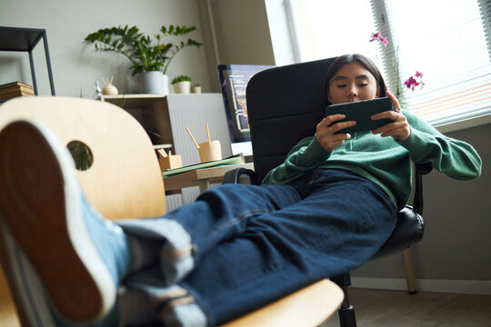 Asian teenage boy reclining in office chair using smartphone with legs stretched out on desk, working in modern office environment with digital devices and plants visible in background