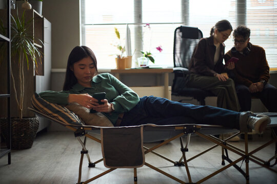 Young Asian woman lying on folding cot using smartphone in modern office, while two young Asian coworkers sitting together in background discussing information on mobile device