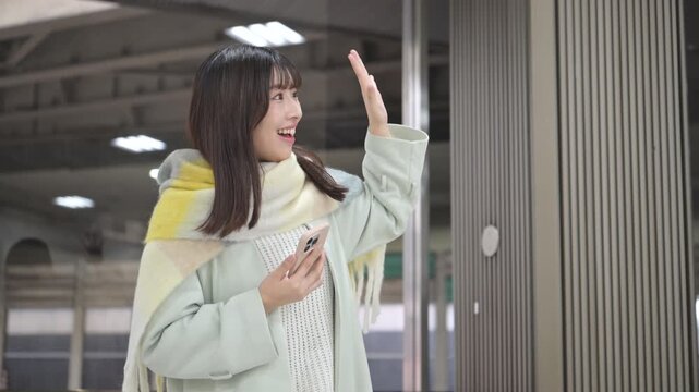 冬の年末年始の帰省、駅のホームやバスターミナルなどでスマホを持つかわいいストールを巻いた女性、手を振るバイバイのポーズ動画