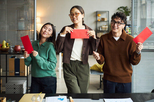 Three young adult Asian men and women standing in modern office holding red envelopes, smiling and looking at camera, business documents and laptop visible on desk in foreground - Powered by Adobe