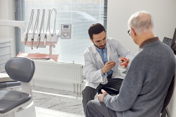 Middle Eastern male dentist explaining dental procedure to senior Caucasian man using dental model in modern dental office, both seated and engaged in consultation