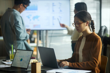 Young Asian businessmen and businesswomen working in modern office, young Asian woman using laptop at desk while two young Asian colleagues discussing data on digital screen in background