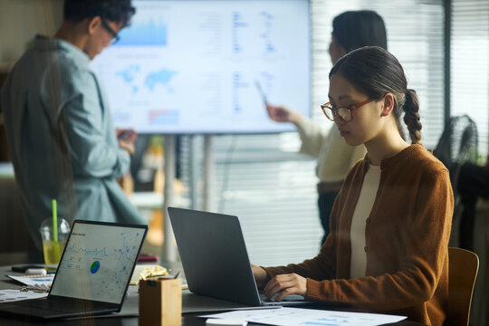 Young Asian businessmen and businesswomen working in modern office, young Asian woman using laptop at desk while two young Asian colleagues discussing data on digital screen in background