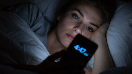 Woman in bed checking her phone at 4:47 AM, unable to sleep due to insomnia, anxiety, or addiction from blue light exposure.