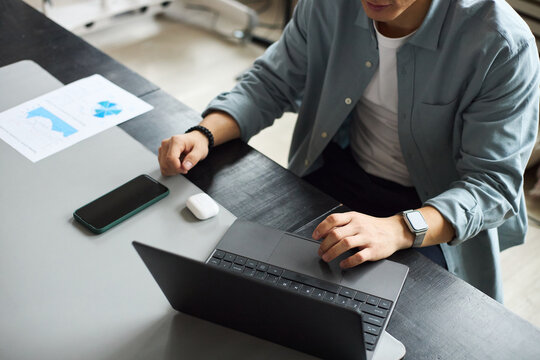 Young Asian man working on laptop at office desk, using wireless earphones and smartphone, analyzing business documents with charts, wearing smartwatch, focused on professional tasks