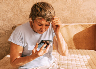 An elderly woman wearing glasses was intently immersed in her smartphone while sitting on a sofa in a sun-drenched. Digital Phone Addiction in Older Women, Screen Time Overload, and Modern Technology